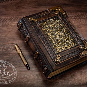 May include: A handcrafted leather journal with ornate gold accents and a matching pen. The journal features a detailed embossed design on the cover and corner protectors. The pen is gold-toned with black ink. The journal is a unique accessory.
