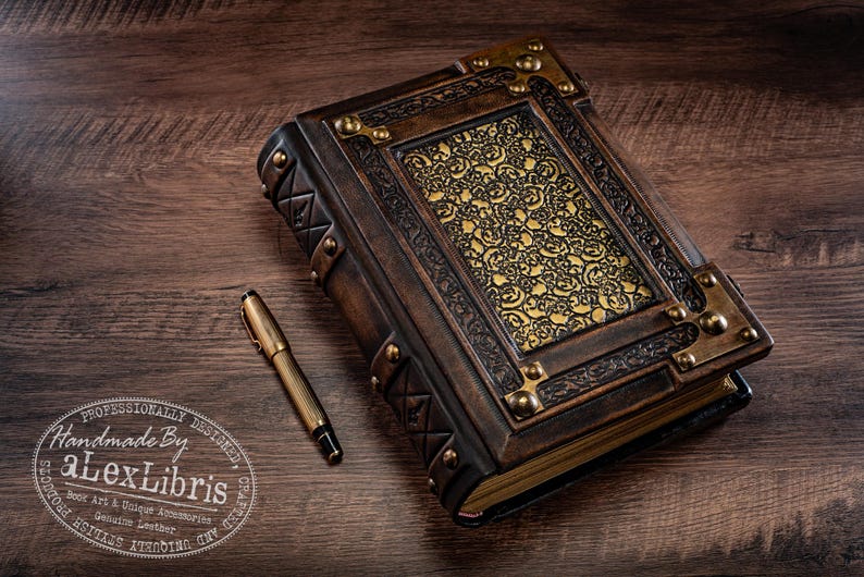 May include: A handcrafted leather journal with ornate gold accents and a matching pen. The journal features a detailed embossed design on the cover and corner protectors. The pen is gold-toned with black ink. The journal is a unique accessory.