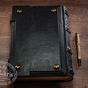 Op de afbeelding: Een groot, zwart lederen boek met gouden accenten en een bijpassende pen. Het boek heeft decoratieve metalen studs en een getextureerd oppervlak. De pen is goudkleurig met zwarte details. Een uniek accessoire.