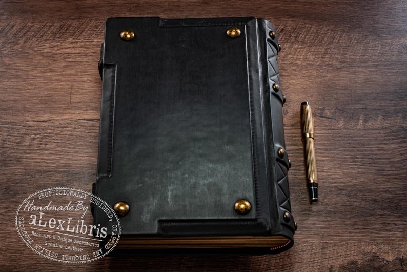 Op de afbeelding: Een groot, zwart lederen boek met gouden accenten en een bijpassende pen. Het boek heeft decoratieve metalen studs en een getextureerd oppervlak. De pen is goudkleurig met zwarte details. Een uniek accessoire.
