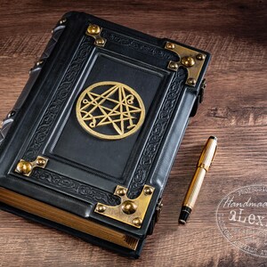 Op de afbeelding: Een zwart leren notitieboek met een gouden embleem en hoekaccenten. Een gouden pen ligt naast het boek op een houten oppervlak. Het notitieboek heeft een sluiting.