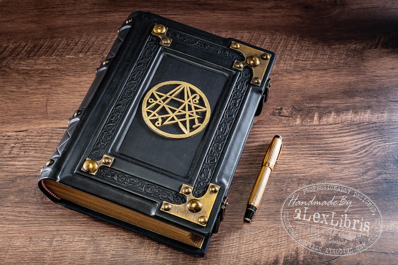 Op de afbeelding: Een zwart leren notitieboek met een gouden embleem en hoekaccenten. Een gouden pen ligt naast het boek op een houten oppervlak. Het notitieboek heeft een sluiting.