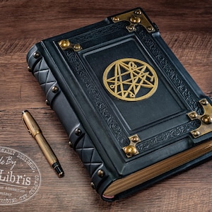 Op de afbeelding: Een donkerblauw leren boek met gouden details en een gouden embleem op de kaft. Een gouden pen ligt naast het boek op een houten oppervlak. Het boek heeft een decoratief ontwerp en is gelabeld "Handmade By aLexLibris".