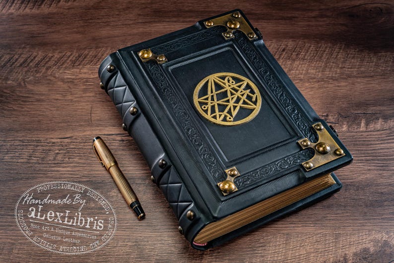 Op de afbeelding: Een donkerblauw leren boek met gouden details en een gouden embleem op de kaft. Een gouden pen ligt naast het boek op een houten oppervlak. Het boek heeft een decoratief ontwerp en is gelabeld "Handmade By aLexLibris".