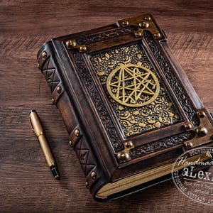 May include: A handcrafted, leather-bound journal with a brass pentagram design. The brown cover features ornate gold accents and metal corner protectors. A gold pen rests beside the book on a wooden surface. The journal is a unique piece of book art.
