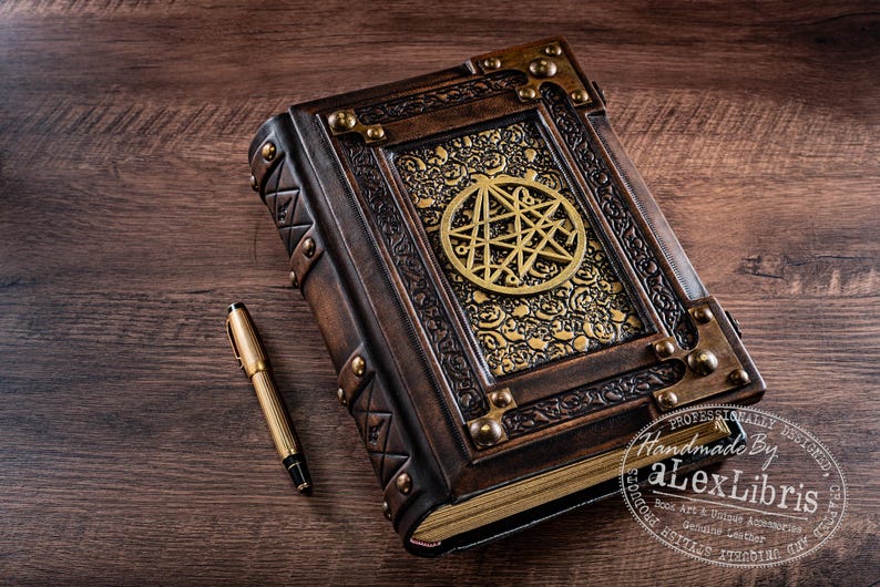 May include: A handcrafted, leather-bound journal with a brass pentagram design. The brown cover features ornate gold accents and metal corner protectors. A gold pen rests beside the book on a wooden surface. The journal is a unique piece of book art.
