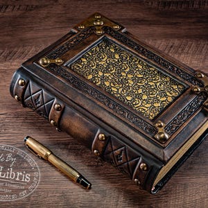 May include: A handcrafted leather journal with ornate gold accents and a decorative gold pattern. A gold pen rests beside the journal. The journal is embossed with geometric designs and metal corner pieces.