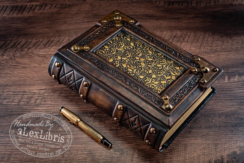 May include: A handcrafted leather journal with ornate gold accents and a decorative gold pattern. A gold pen rests beside the journal. The journal is embossed with geometric designs and metal corner pieces.