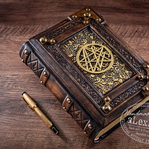 May include: A handcrafted, leather-bound book with a gold pentagram design on the cover. The book is brown with gold accents and metal corner protectors. A gold pen rests beside the book on a wooden surface.