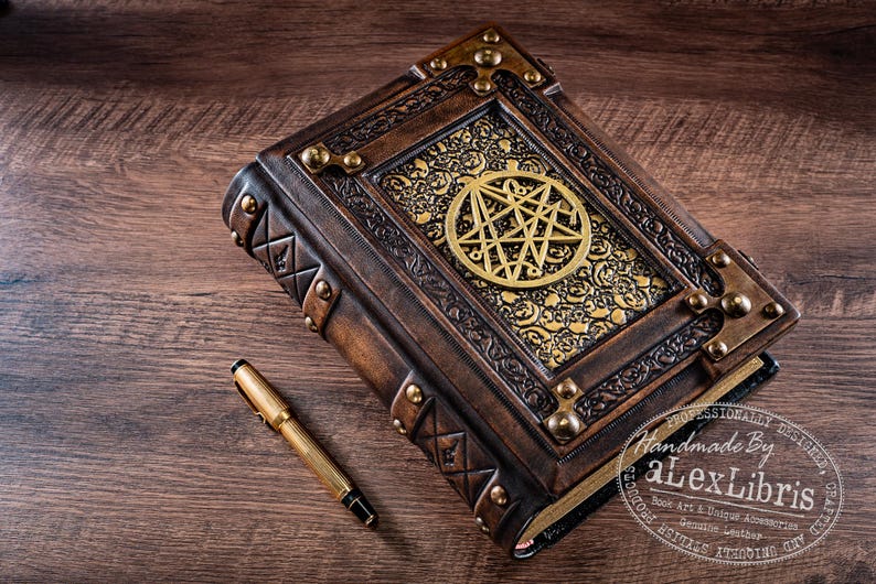 May include: A handcrafted, leather-bound book with a gold pentagram design on the cover. The book is brown with gold accents and metal corner protectors. A gold pen rests beside the book on a wooden surface.