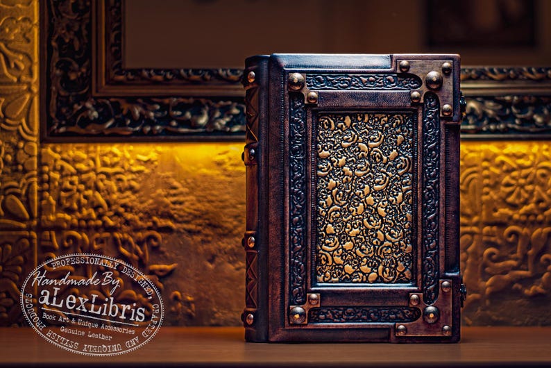 May include: A handcrafted leather book with intricate embossed floral patterns in gold and brown. The book has a decorative frame and metal accents. The text "Handmade By aLexLibris" is visible on the image.