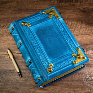Könnte beinhalten: Ein leuchtend blaues, in Leder gebundenes Tagebuch mit verzierten Prägerändern und polierten Messing-Eckschützern. Das Buch ist mit Messingnieten entlang des Buchrückens und der Kanten befestigt. Ein goldfarbener Füllfederhalter liegt daneben.