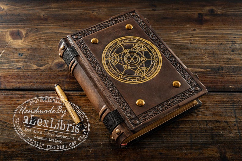 K&ouml;nnte beinhalten: Ein braunes, ledergebundenes Buch mit einem goldenen geometrischen Design auf dem Einband. Das Buch hat dekorative Metallnieten und einen schwarzen R&uuml;cken. Ein goldener Stift und ein Logo mit der Aufschrift "Handmade By aLexLibris" sind ebenfalls vorhanden.