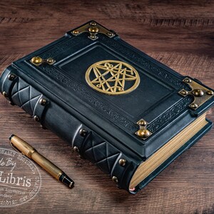 Op de afbeelding: Een donkerblauw, ledergebonden boek met gouden accenten en een gouden pentagram-embleem. Een gouden pen ligt naast het boek op een houten oppervlak. Het boek heeft decoratieve metalen hoekstukken en een gewatteerd patroon.