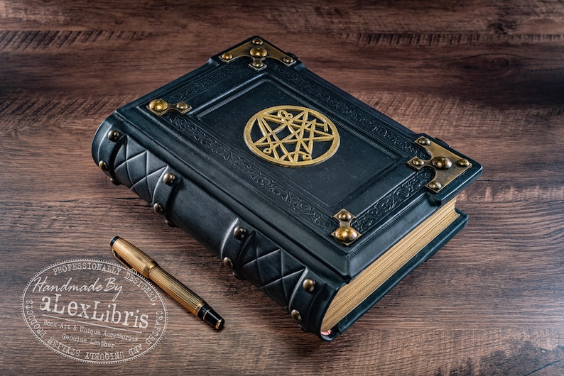 Op de afbeelding: Een donkerblauw, ledergebonden boek met gouden accenten en een gouden pentagram-embleem. Een gouden pen ligt naast het boek op een houten oppervlak. Het boek heeft decoratieve metalen hoekstukken en een gewatteerd patroon.