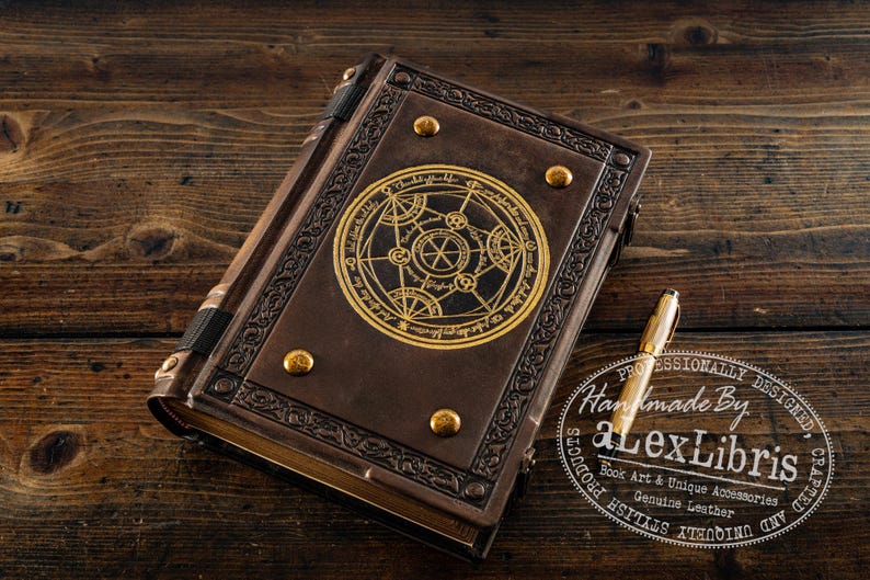 K&ouml;nnte beinhalten: Ein braunes, ledergebundenes Notizbuch mit einem goldgepr&auml;gten Design auf dem Einband, das ein kreisf&ouml;rmiges Symbol zeigt. Goldene Akzente und ein goldener Stift sind sichtbar. Das Notizbuch liegt auf einer Holzoberfl&auml;che.