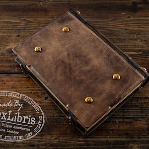 K&ouml;nnte beinhalten: Ein handgefertigtes, braunes Lederjournal mit Messingakzenten. Der Einband ist gealtert und strukturiert, mit Messingkn&ouml;pfen befestigt. Das Journal stammt von aLexLibris, bekannt f&uuml;r Buchkunst und einzigartige Accessoires.