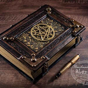 May include: A handcrafted, leather-bound book with a brass pentagram emblem and decorative corner accents. A gold-toned pen rests beside the book. The book's cover features ornate gold filigree. The text "Handmade By aLexLibris" is visible.