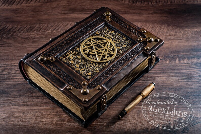 May include: A handcrafted, leather-bound book with a brass pentagram emblem and decorative corner accents. A gold-toned pen rests beside the book. The book's cover features ornate gold filigree. The text "Handmade By aLexLibris" is visible.