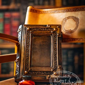 Könnte beinhalten: Ein dunkelbraunes Lederjournal mit geprägtem rechteckigem Rahmen und Metallnieten, begleitet von einem kleinen Glas-Tintenfass mit roter Tinte und einem goldfarbenen Füllfederhalter. Eine Lederrolle mit einem kreisförmigen Emblem ist sichtbar.