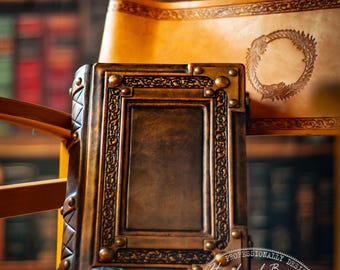 Puede incluir: Un diario de cuero marrón oscuro con un marco rectangular en relieve y tachuelas metálicas, acompañado de un pequeño tintero de cristal con tinta roja y una pluma estilográfica dorada. Se ve un pergamino de cuero con un emblema circular.