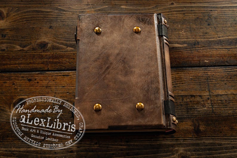 K&ouml;nnte beinhalten: Ein handgefertigtes, braunes Lederjournal mit Messingakzenten. Das geschlossene Journal hat vier Messingkn&ouml;pfe auf dem Einband und eine detaillierte R&uuml;ckenpartie. Das Bild enth&auml;lt ein Logo mit der Aufschrift "Handmade By Alex Libris".