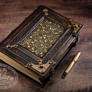 May include: A handcrafted, leather-bound journal with ornate brass accents and a gold-toned, patterned center panel. A gold pen rests beside the book on a dark wood surface. The journal is labeled "aLexLibris".