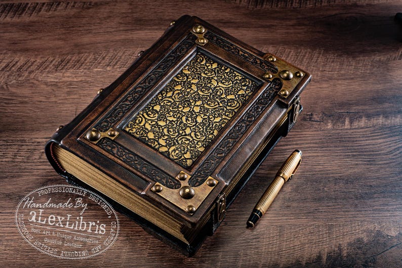 May include: A handcrafted, leather-bound journal with ornate brass accents and a gold-toned, patterned center panel. A gold pen rests beside the book on a dark wood surface. The journal is labeled "aLexLibris".