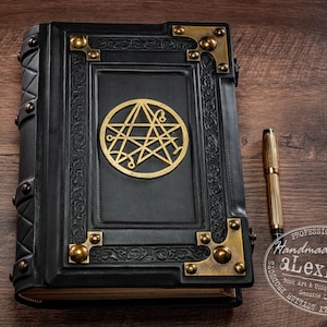Op de afbeelding: Een zwart leren boek met gouden details en een gouden embleem op de kaft. Een gouden pen ligt naast het boek op een houten oppervlak. Het boek heeft decoratieve hoekstukken en een geniete rug.