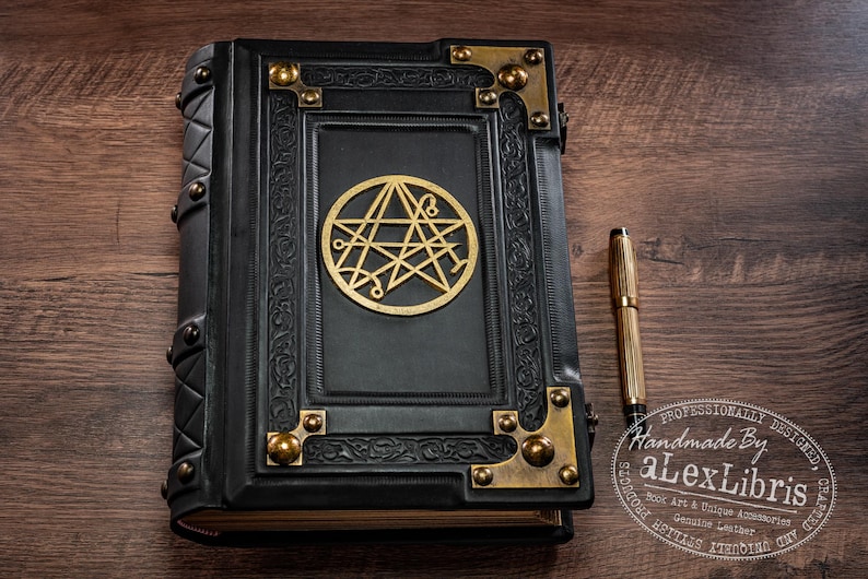 Op de afbeelding: Een zwart leren boek met gouden details en een gouden embleem op de kaft. Een gouden pen ligt naast het boek op een houten oppervlak. Het boek heeft decoratieve hoekstukken en een geniete rug.