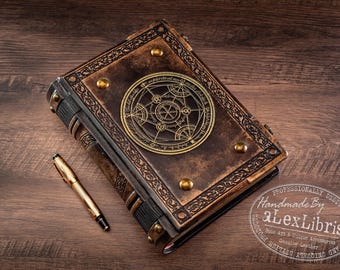 Puede incluir: Un diario encuadernado en cuero marrón con un intrincado emblema circular de metal dorado en la portada, tachuelas decorativas de metal en las esquinas y patrones de borde en relieve. Un bolígrafo de metal dorado se sitúa junto al libro.