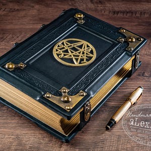 Op de afbeelding: Een donkerblauw leren boek met goud vergulde pagina's en messing hoekaccenten. Een gouden pen ligt naast het boek. Het boek heeft een gouden embleem op de kaft. Het boek ligt op een houten oppervlak.