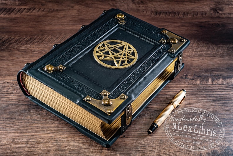 Op de afbeelding: Een donkerblauw leren boek met goud vergulde pagina's en messing hoekaccenten. Een gouden pen ligt naast het boek. Het boek heeft een gouden embleem op de kaft. Het boek ligt op een houten oppervlak.
