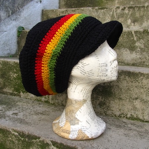 Könnte beinhalten: Ein schwarzer Häkelhut mit Visor, mit einem leuchtenden Streifen aus Rot, Gelb und Grün. Der Hut wird auf einem Schaufensterkopf präsentiert. Das Design des Hutes erinnert an die Rastafari-Kultur.