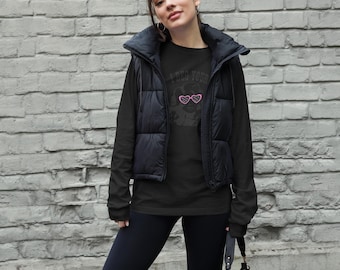 Könnte beinhalten: Ein schwarzes Langarmshirt mit einer Grafik einer Person, die eine rosa herzförmige Sonnenbrille trägt, und dem Text I BEG YOUR PARDON, kombiniert mit einer schwarzen Steppweste, einer schwarzen Leggings und einer schwarzen Umhängetasche.