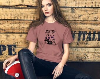 Könnte beinhalten: Ein altrosa kurzärmeliges T-Shirt mit einer schwarzen grafischen Illustration einer Person mit welligem Haar, die herzförmige Brillen trägt, wobei der Text "I BEG YOUR" in Schwarz über der Illustration und eine Signatur darunter gedruckt ist.