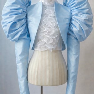May include: A light blue bolero jacket with dramatic, puffed sleeves and long, fitted sleeves. The jacket is displayed on a mannequin over a white ruffled blouse. The overall style is elegant and vintage-inspired.