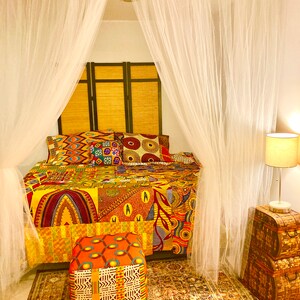 May include: A bed with a colorful African print quilt and matching pillows, a white mosquito net, and a small ottoman with a similar print. The bed is in a room with a wooden headboard and a wooden side table with a lamp.