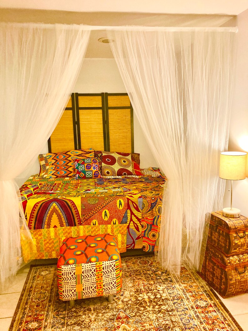 May include: A bed with a colorful African print quilt and matching pillows, a white mosquito net, and a small ottoman with a similar print. The bed is in a room with a wooden headboard and a wooden side table with a lamp.