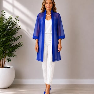 May include: A woman wearing a royal blue sheer duster coat over a white top and pants. The coat has three-quarter sleeves and a vertical stripe down the front. She is wearing blue heels and a pearl necklace. A potted plant is in the background.