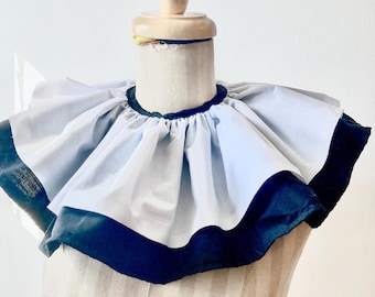 May include: A white ruffled collar with a black velvet trim. The collar is on a dress form.
