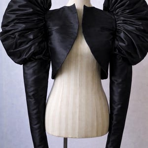 May include: Black bolero jacket with dramatic, oversized puffed sleeves. The jacket is displayed on a mannequin, showcasing its unique design. The sleeves are long and fitted, contrasting with the voluminous shoulders.