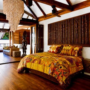 Könnte beinhalten: Ein Schlafzimmer mit einem Bett, das mit einer farbenfrohen Patchwork-Tagesdecke in Gelb-, Orange- und Rottönen bezogen ist. Über dem Bett hängt ein großes Wandpaneel aus dunklem Holz mit geschnitzten Figuren. Ein mehrstufiger Muschel-Kronleuchter.