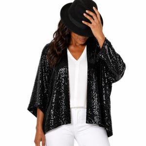 May include: A black sequin jacket with wide sleeves, worn over a white V-neck top and white pants. A black fedora hat is worn on the head. The jacket is covered in small, reflective sequins.
