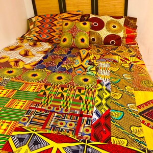 May include: A colorful patchwork quilt with a variety of African print fabrics. The quilt is made up of squares and rectangles of different colors and patterns, including yellow, red, green, blue, and black. The quilt is laid out on a bed with three pillows.