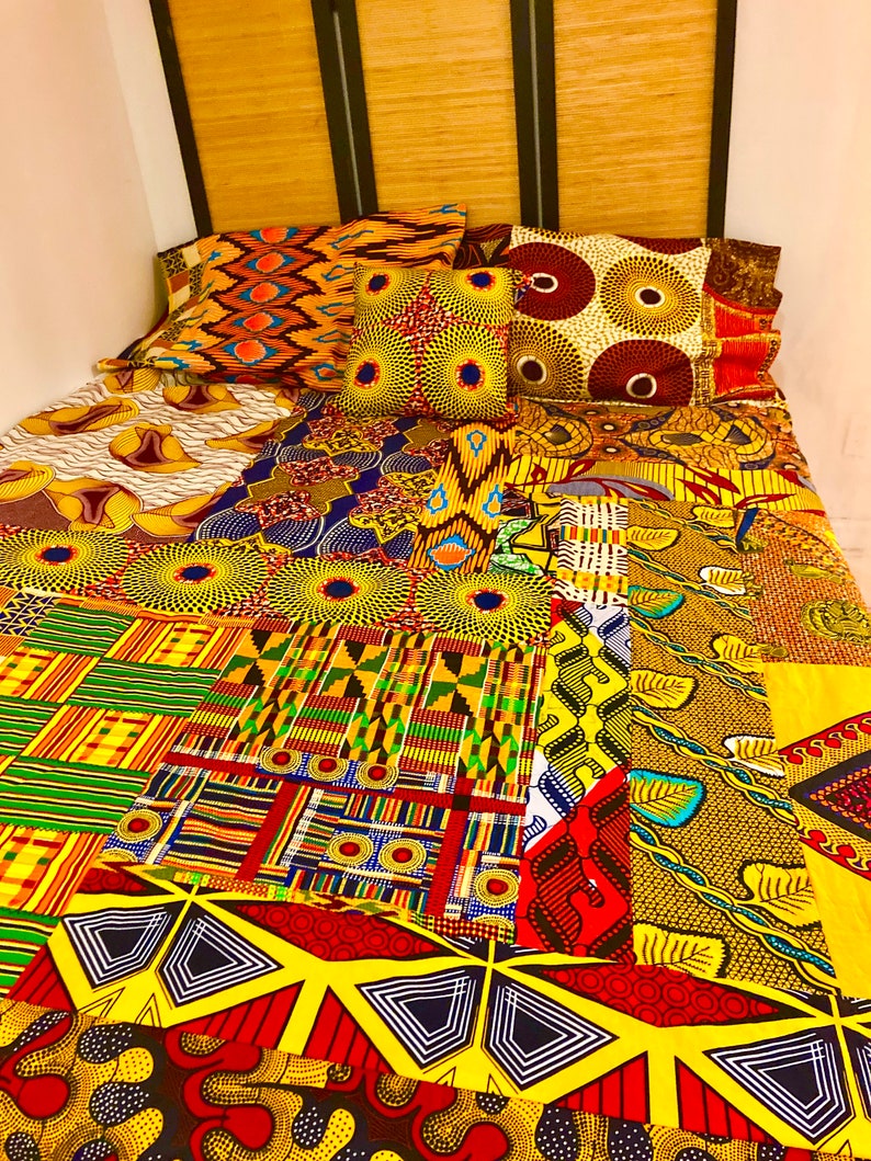 May include: A colorful patchwork quilt with a variety of African print fabrics. The quilt is made up of squares and rectangles of different colors and patterns, including yellow, red, green, blue, and black. The quilt is laid out on a bed with three pillows.