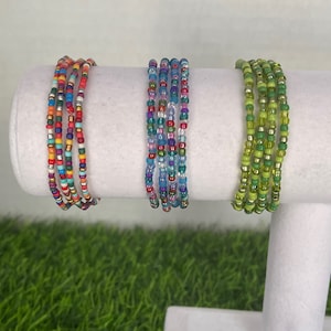 May include: Three colorful beaded bracelets on a white display stand. The bracelets are made of small, colorful beads and are stacked on top of each other. The bracelets are on a white display stand against a white background.