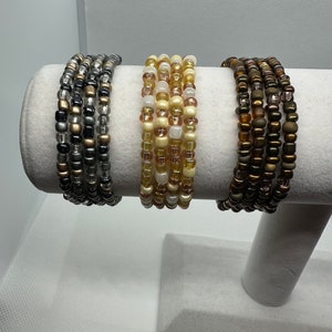 May include: Three beaded bracelets on a white display stand. The bracelets are made of small, colorful beads in shades of black, gold, cream, and brown.