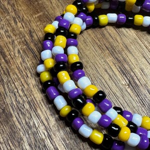 May include: Close-up of three strands of beaded jewelry. The beads are in a pattern of yellow, black, white, and purple. The jewelry is on a wooden surface.