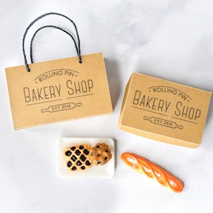 May include: Two brown cardboard boxes with the text "Rolling Pin Bakery Shop Est. 2019" printed on them. One box has a handle. A white plate with a chocolate chip cookie and a waffle with chocolate filling. A long, orange baguette.